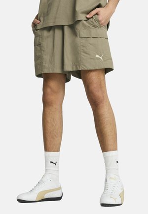 Khaki cargo shorts med to sidelommer, løs pasform, ensartet tekstur og et lille hvidt Puma-logo. Båret med hvide og guld sneakers og stribede sokker.
