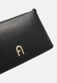 Portafoglio in pelle nera con un'accentatura decorativa in metallo dorato a forma di "A", con chiusura a zip lungo il bordo superiore.