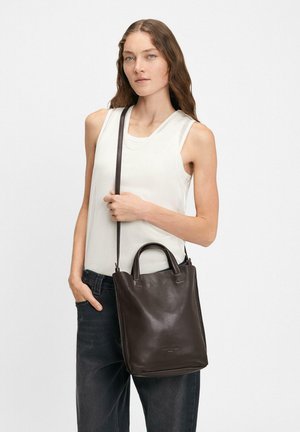 Brązowa skórzana torba tote o wyraźnej strukturze, z krótkimi uchwytami i długim paskiem na ramię, noszona przez modelkę na jednolitym tle.