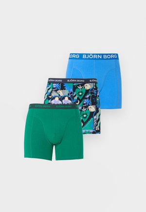 Νεαρός μαύρος άνδρας στέκεται ξυπόλητος πάνω σε χαλί, φορώντας πράσινα ανδρικά εσώρουχα τύπου μπόξερ, κοιτάζοντας πλάγια προς έναν απλό λευκό τοίχο στο φόντο.