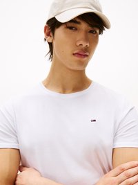 Junger Mann, der eine weiße Mütze und ein weißes T-Shirt mit einem kleinen rot-weiß-blauen Logo trägt, steht mit verschränkten Armen vor einem weißen Hintergrund.