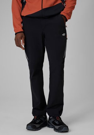 Osoba ubrana w czarne outdoorowe spodnie marki 4F i czarne buty trekkingowe z pomarańczowo-czarną kurtką, jedną ręką w kieszeni, stojąca na szarym tle.