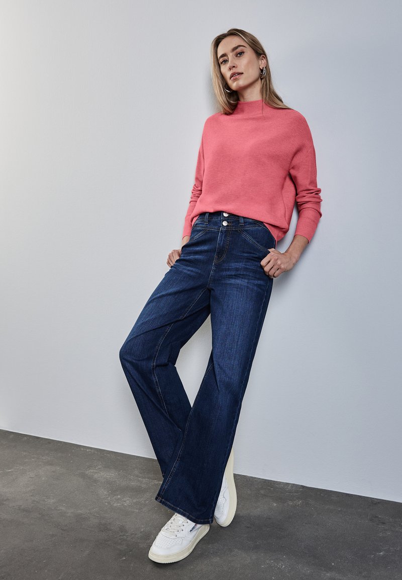 Un pull en tricot rose avec un col montant, associé à un jean en denim large bleu foncé et des baskets blanches, sur un fond neutre.