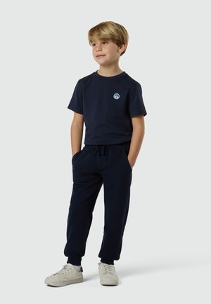 Junge mit hellbraunem Haar, der ein marineblaues T-Shirt und Hose trägt, mit den Händen in den Taschen steht und weiße Turnschuhe auf hellem Hintergrund trägt.