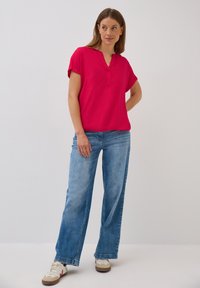Blouse fuchsia à manches courtes avec un col en V et une patte de boutonnage, associée à un jean large bleu clair. Tissu lisse et coupe décontractée.