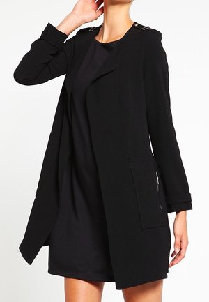 Frau trägt einen schwarzen, langärmeligen Mantel mit offener Vorderseite über einem schwarzen Kleid, steht mit einer Hand am Hals und der anderen entspannt an der Seite.