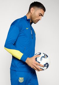 Μπλε αθλητικό μπουφάν με κίτρινες λεπτομέρειες, γιακά με φερμουάρ και λογότυπο. Μπλε παντελόνια που ταιριάζουν με έμβλημα. Άτομο που κρατά μια μπάλα ποδοσφαίρου.