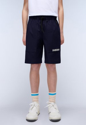 Pantalones cortos de color azul marino con cinturilla elástica y cordón. Presentan bolsillos laterales y un parche de logo. Combinados con zapatillas blancas y calcetines a rayas beige.