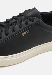 Čierne kožené tenisky s textúrovaným povrchom, šnurovací dizajn a zlaté logo „BOSS“. Obsahujú bielu gumovú podrážku s čistými, minimalistickými líniami.