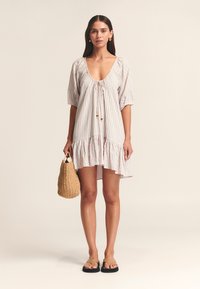 Gestreepte jurk met een V-hals, korte pofmouwen, gerafelde zoom en een trekkoorddetail. Model houdt een geweven strozak vast en draagt platte sandalen.