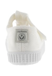 Witte canvas sneaker met een getextureerde rubberen zool, voorzien van een stoffen lus bij de hak en een grijze logopatch op de achterkant.