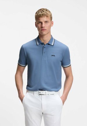 Mężczyzna w niebieskiej koszulce polo z czarno-białym paskowanym kołnierzem i rękawami, założonej do białych spodni z białym paskiem, ręce w kieszeniach.