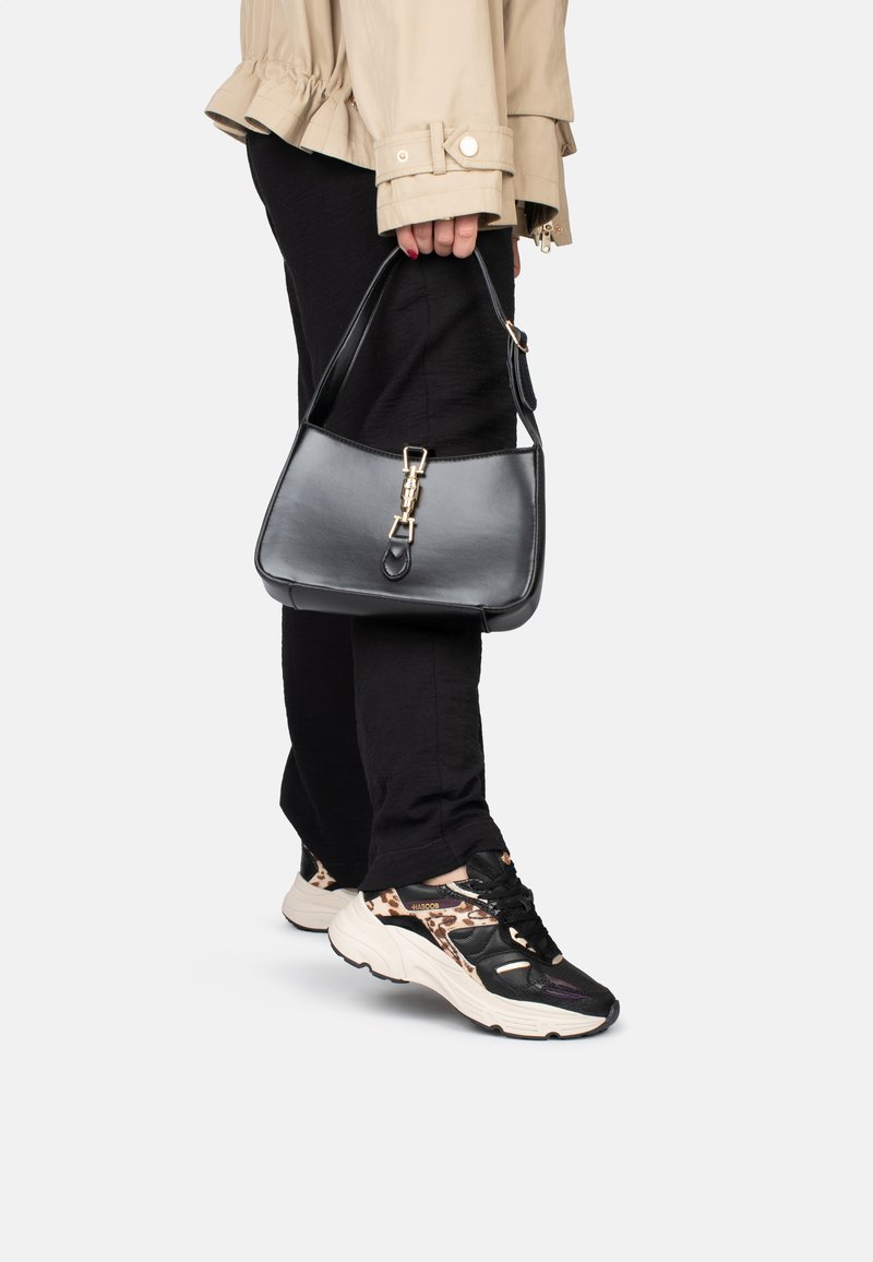 Borsa a mano in pelle nera con una forma strutturata, dotata di una chiusura dorata e una lunga tracolla. Indossata con pantaloni neri e sneakers con motivo.