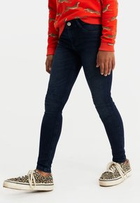 Tmavě modré skinny džíny s hladkou texturou, které mají klasický pětikapsový design a přední zapínání na knoflík, nošeny s teniskami se zvířecím potiskem.