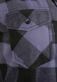 Tkanina s kostkovaným vzorem v černé a fialové barvě, která obsahuje náprsní kapsu s černým knoflíkem. Texturovaný povrch s viditelnými detaily šití.