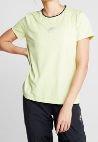 Osoba v světle zeleném sítovaném tričku Nike Air a černých kalhotách Nike Air, stojící s jednou rukou na boku a druhou uvolněnou.