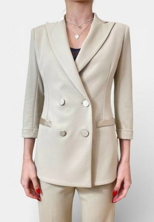 Donna che indossa un blazer doppiopetto beige con maniche a tre quarti e pantaloni abbinati, che mostra collane argento a strati e smalto per unghie rosso.