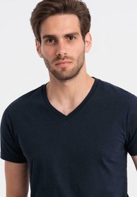 Giovane uomo con capelli corti castani e barba, indossa una semplice maglietta scura a V blu navy su uno sfondo chiaro.