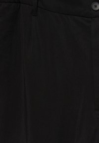 Gros plan d'un pantalon en tissu noir montrant un bouton, la ceinture, une passants de ceinture et des détails de couture.
