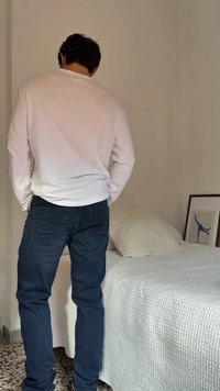 Άτομο που φοράει λευκό μπλουζάκι μακρυμάνικο και μπλε τζιν στέκεται με την πλάτη στον τοίχο δίπλα σε κρεβάτι με λευκή υφασμάτινη κουβέρτα και μαξιλάρι.