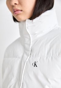 Vit pufferjacka med hög krage, slät textur och logotypen "cK" på framsidan. Sytt detaljarbete längs dragkedjan.
