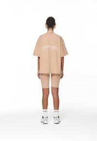 Kvinna med uppsatt hår som bär en beige oversized t-shirt med text och matchande shorts, vita strumpor och sneakers, stående vänd bort.