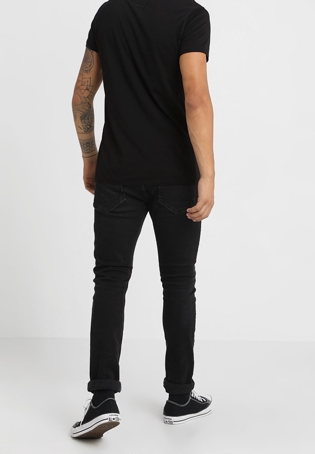 Sort t-shirt med korte ærmer, sorte slim-fit jeans og sorte sneakers med hvide detaljer. Mandens ryg er synlig, hvilket viser tatoveringer på venstre arm.