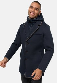 Marineblå frakke med en tætsiddende pasform, der har en dobbeltsidet hætte, kontrasterende syninger og knaplukninger. Tekstureret stof og sidelommer.