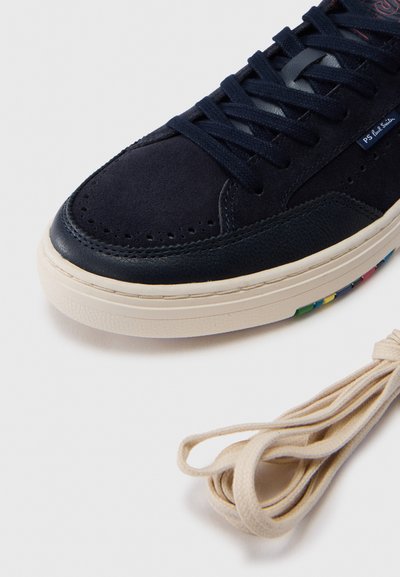 Κοντινό πλάνο  αθλητικού παπουτσιού σκούρου μπλε με υφές από σουέντ και δέρμα, λευκή σόλα και επιπλέον μπεζ κορδόνια σε λευκό φόντο.