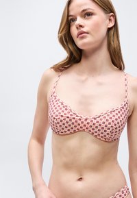 Νεαρή γυναίκα που φοράει μπικίνι πάνω μέρος με ροζ και κόκκινο μοτίβο και λεπτές τιράντες, στέκεται μπροστά σε απλό ανοιχτό γκρι φόντο.