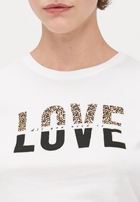 Bílý tričko s nápisem "LOVE" v černých a leopardích písmenech. Pod ním je v menším černém textu fráze "Všechno, co potřebujete, je". Měkký bavlněný materiál.