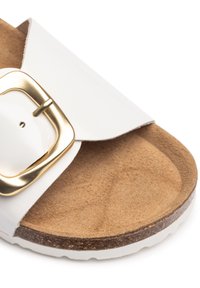 Weiße Slide-Sandale mit einem breiten Riemen und goldfarbenem Schnallenverschluss, ausgestattet mit einem strukturierten Korkfußbett und einer stabilen weißen Gummisohle.