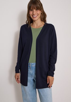 Femme souriante portant un cardigan ouvert bleu marine foncé, un haut vert et un jean en denim bleu clair sur un fond clair uni.