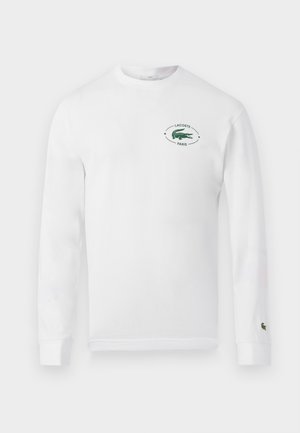 Deux mannequins portent des chemises blanches à manches longues et des pantalons de jogging noirs Lacoste avec des baskets blanches, debout devant un fond blanc uni.