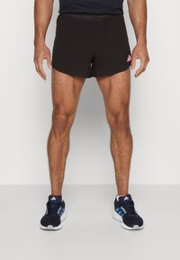 Svarta träningsshorts med perforerad textur och rosa logotyp på vänster sida, i kombination med blå och vita träningsskor.