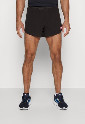 Pantalones cortos deportivos negros con una textura perforada y un logo rosa en el lado izquierdo, combinados con zapatillas deportivas azules y blancas.