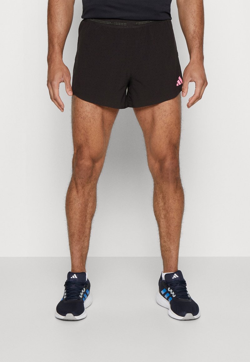 Svarta träningsshorts med perforerad textur och rosa logotyp på vänster sida, i kombination med blå och vita träningsskor.