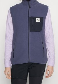 Eine Frau trägt eine lila Fleeceweste mit Reißverschluss über einem helllila Langarmshirt und schwarzen Hosen mit Reißverschlusstaschen und Logo-Details.