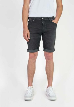 Zwarte denim shorts met een opgerolde zoom, twee voorzakken en een ritssluiting met knoop. Gecombineerd met witte sneakers op een eenvoudige achtergrond.