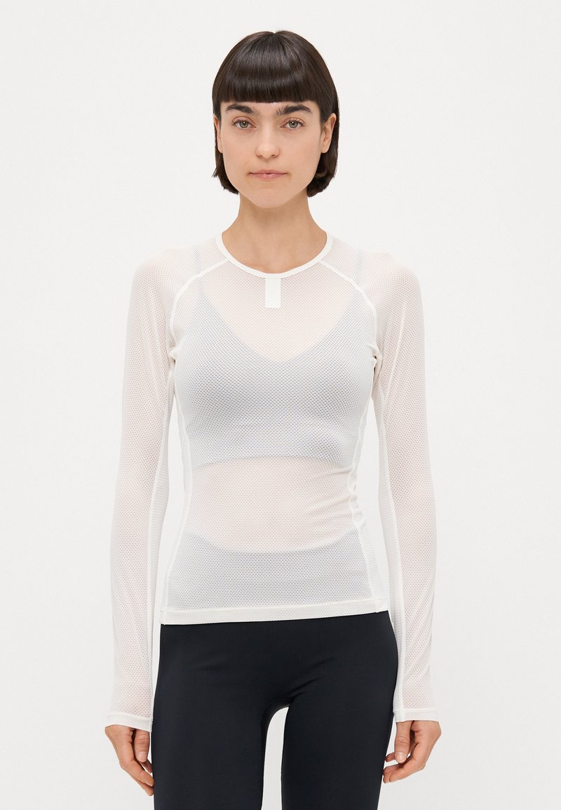 Hvid langærmet mesh-top med rund halsudskæring, der har et struktureret mønster og en tætsiddende pasform, lagdelt over en simpel hvid tanktop.