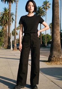 Woman with short black hair wears black t-shirt, wide-leg pants, and sneakers standing on palm tree-lined sidewalk in daytime.