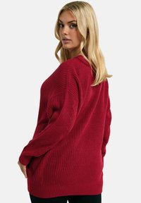 Burgunderfarbener Strickpullover mit lockerer Passform, geripptem Textur, langen Ärmeln und abgesenkten Schultern, kombiniert mit schwarzen Unterteilen.