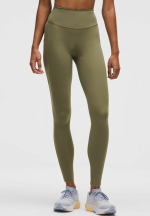 Personne portant un legging taille haute vert olive et des chaussures de sport grises avec des semelles orange, se tenant devant un fond blanc uni.