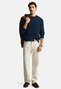 Man draagt een marineblauwe kabelgebreide trui, beige broek, bruine schoenen en houdt een bruine jas over zijn schouder tegen een effen achtergrond.