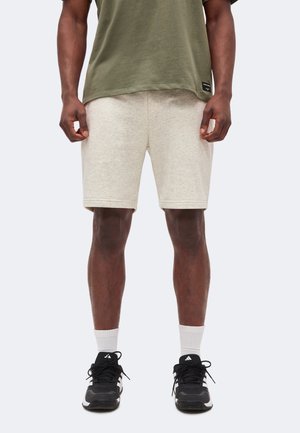 Mand iført beige shorts, olivengrøn T-shirt, hvide sokker og sorte Adidas-sneakers står med hænderne afslappet ned langs siderne.