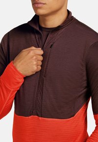 Hombre con un pulóver de bloques de color, con mangas naranjas y cuerpo superior burdeos, desabrochándose parcialmente la cremallera frontal.
