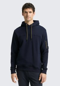 Marineblauwe hoodie met een kangoeroezak, een koord in de capuchon en geribde manchetten. Voorzien van een ritszak op de mouw met gouden accenten. Gelaagde stof.