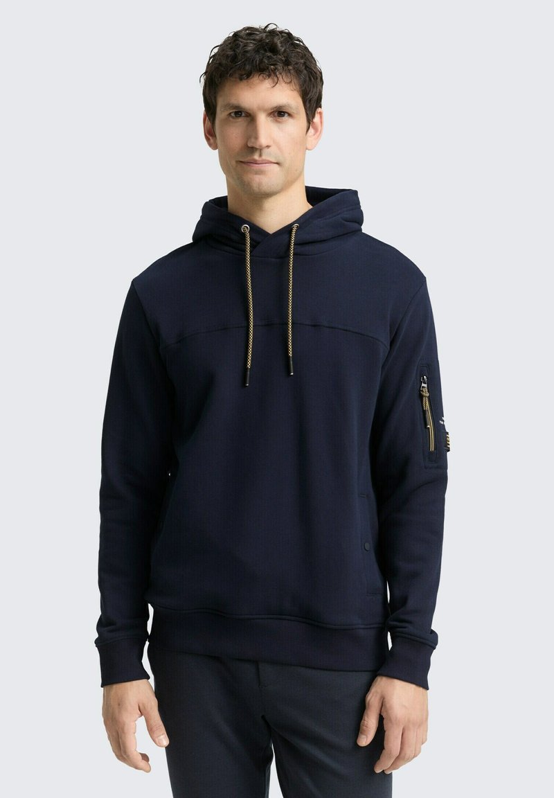 Marineblauwe hoodie met een kangoeroezak, een koord in de capuchon en geribde manchetten. Voorzien van een ritszak op de mouw met gouden accenten. Gelaagde stof.