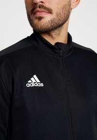 Zwarte ritsjack gemaakt van smooth stof, met een hoge kraag en het witte Adidas-logo op de linkerborst, met zwarte accenten op de schouders.