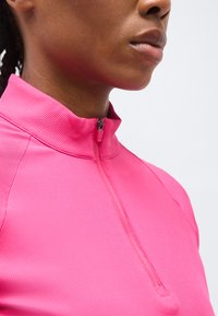 Κοντινό πλάνο του λαιμού και του κάτω μέρους του προσώπου ενός ατόμου που φοράει ένα φωτεινό ροζ κορδωτό μπλουζάκι με φερμουάρ, σε απλό λευκό φόντο.